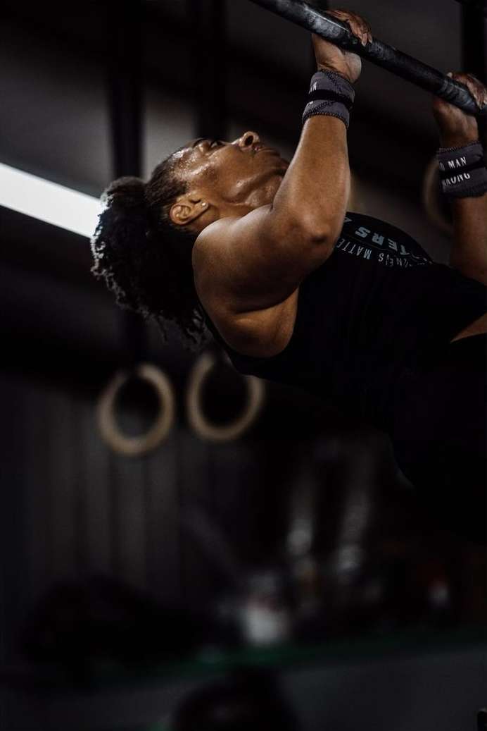 An adult female athlete is hanging from a pull-up bar, wearing wrist wraps and a tank top in a gym with neutral lighting.