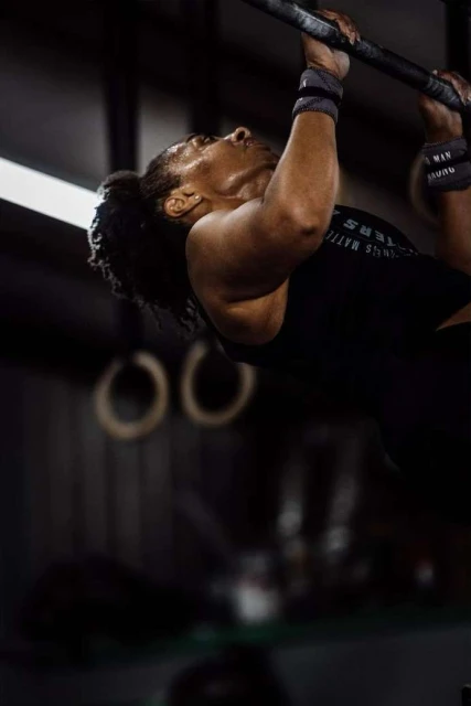 An adult female athlete is hanging from a pull-up bar, wearing wrist wraps and a tank top in a gym with neutral lighting.