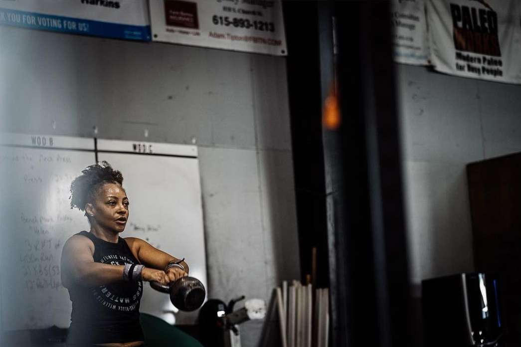 A woman in a fitness tank top works out in a gym, lifting a kettlebell with both hands. The gym has weights and whiteboards.