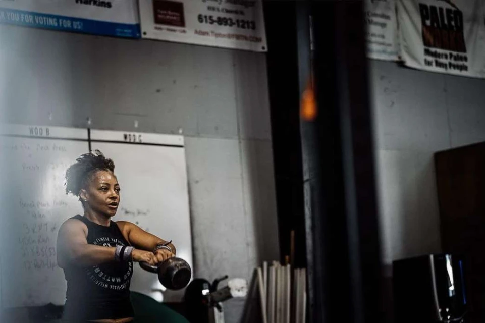 A woman in a fitness tank top works out in a gym, lifting a kettlebell with both hands. The gym has weights and whiteboards.