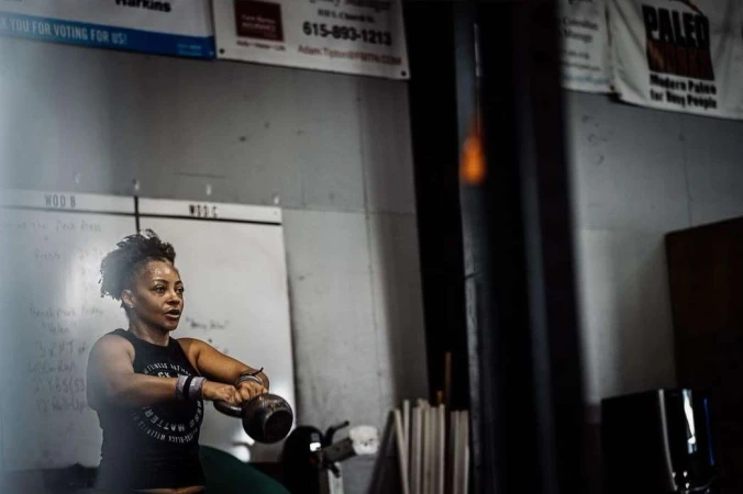 A woman in a fitness tank top works out in a gym, lifting a kettlebell with both hands. The gym has weights and whiteboards.