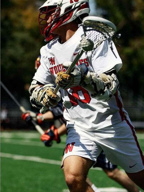 A lacrosse player in uniform and helmet holds a lacrosse stick on a green field, in a medium shot displaying equipment and action.