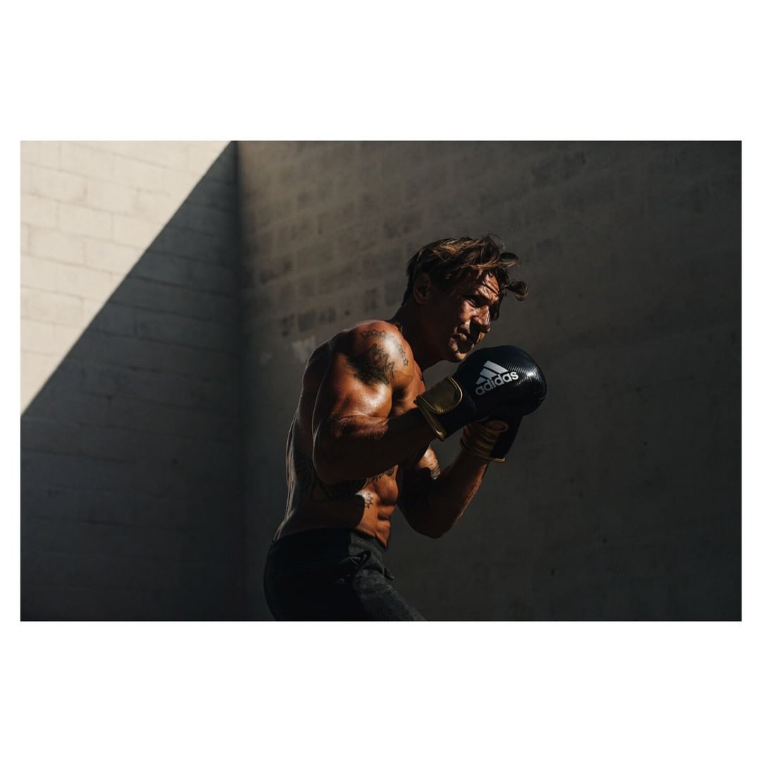 Muscular adult male boxer in black shorts and Adidas boxing gloves, posing in fighting stance in an outdoor urban environment.
