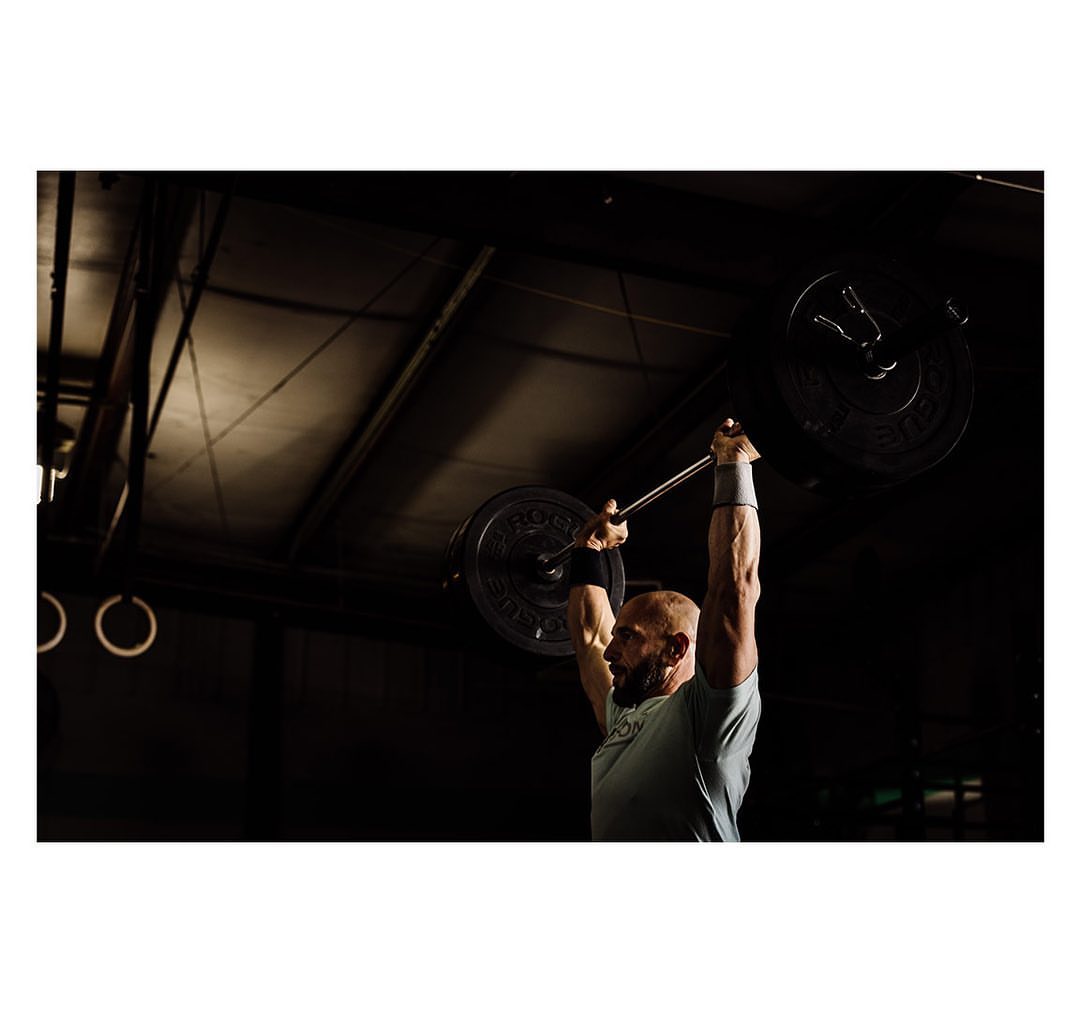 Athletic man lifts barbell overhead in a gym, spotlight illuminates his muscular form, displaying strength and determination in fitness setting.