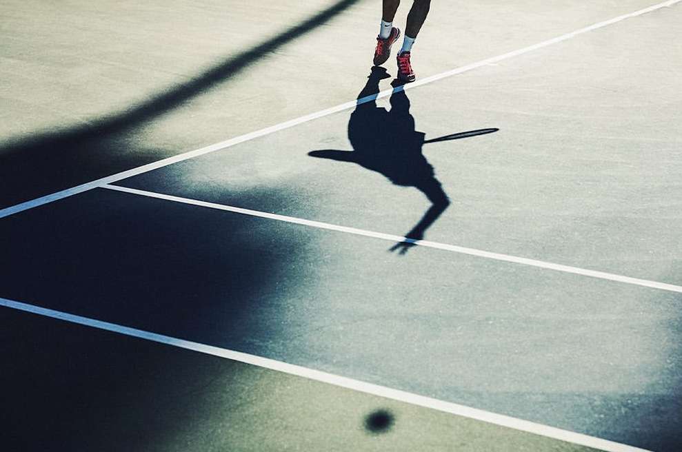 Legs of tennis player jumping in the air on a blue and green court with white lines and the dark shadow of player and tennis racket.