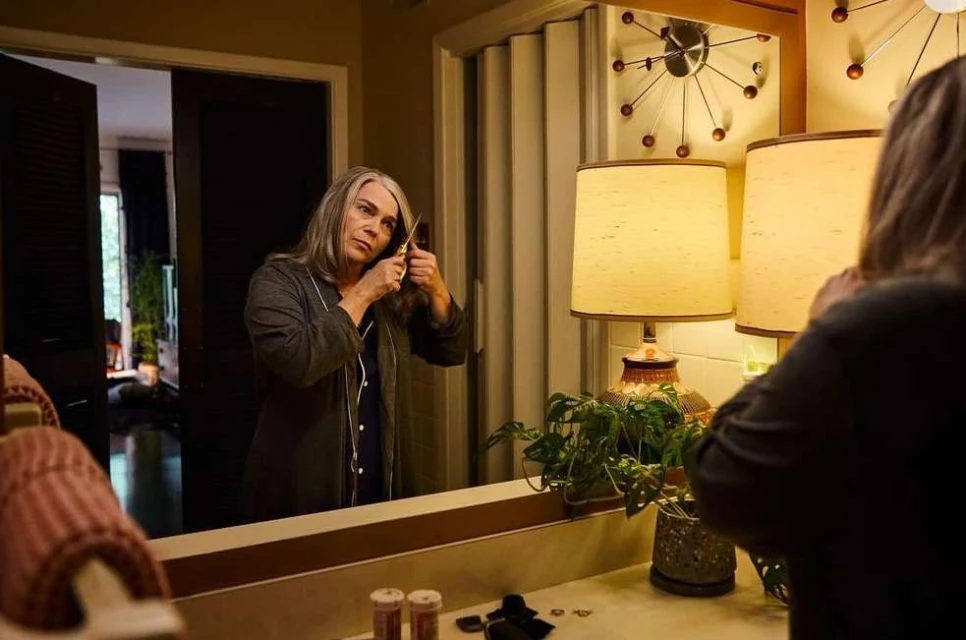 Mature adult woman with grey hair is seen in a bathroom, holding scissors and trimming her own hair in front of a mirror.