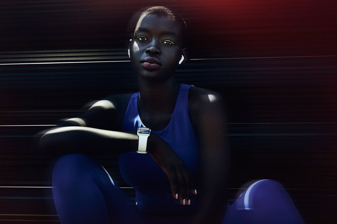 Portrait of a dark-skinned woman wearing a blue top and leggings, white wireless earbuds, and a white smartwatch, sitting outdoors.