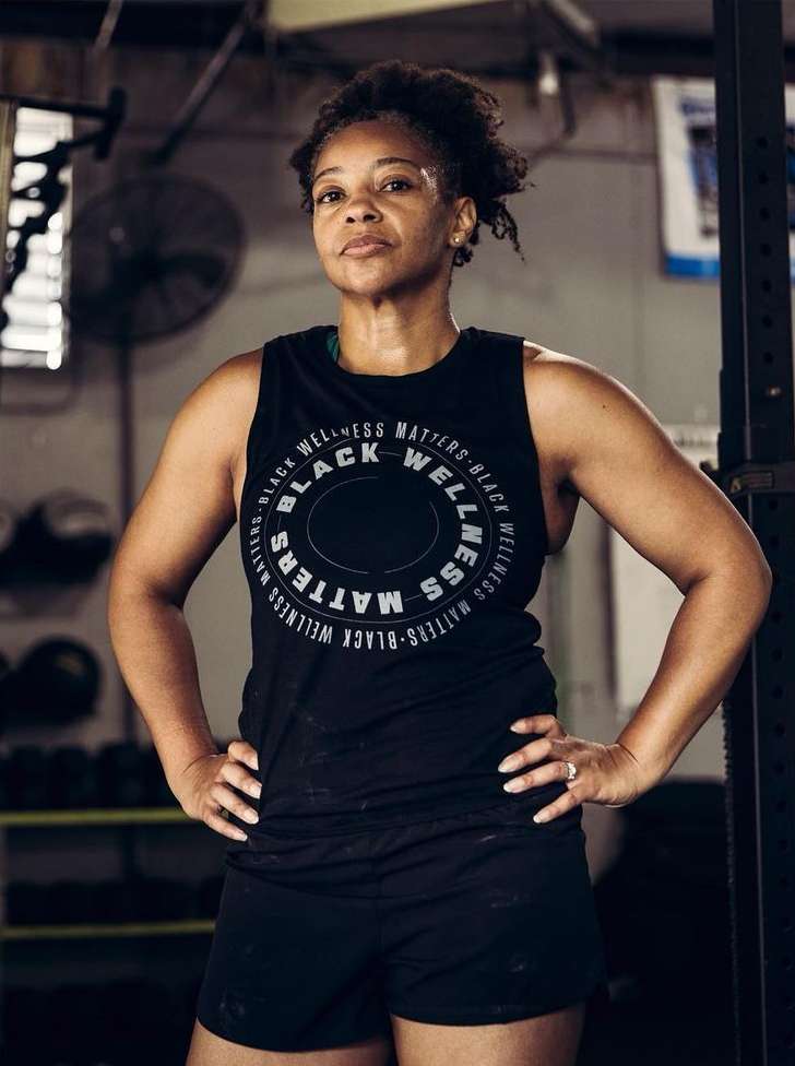 Adult woman in a black tank top with 'Black Wellness Matters' print, posing in a gym with hands on hips, looking forward confidently.