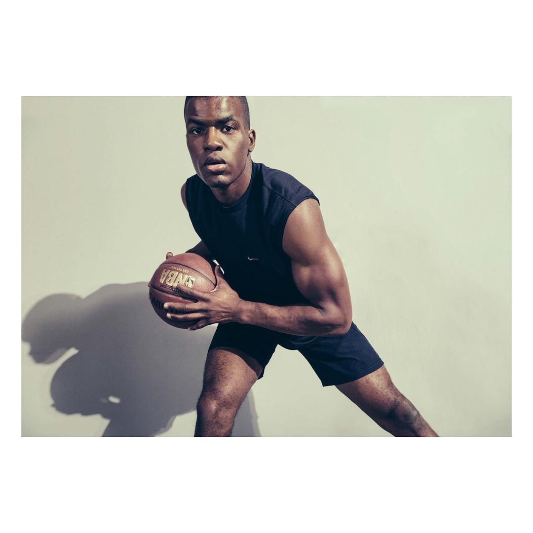 Athletic Black man in sportswear dribbles a basketball indoors against a neutral background, captured in a medium shot with studio lighting.
