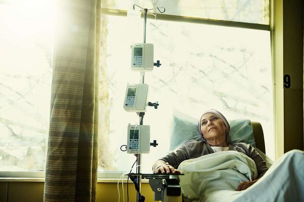 Elderly woman rests in a hospital bed with IV equipment visible. Natural light streams through a window with closed beige curtains.