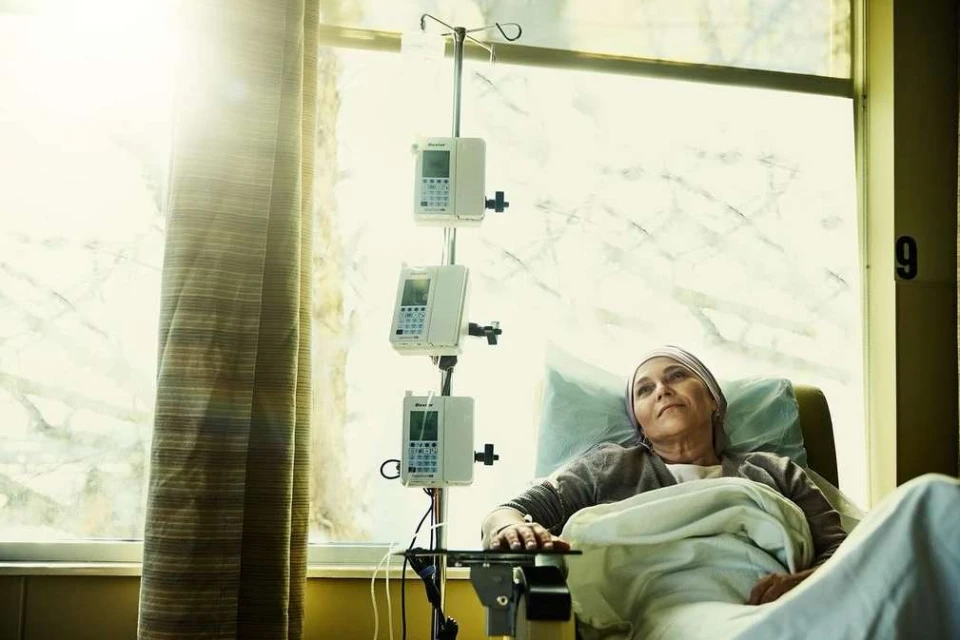 Elderly woman rests in a hospital bed with IV equipment visible. Natural light streams through a window with closed beige curtains.