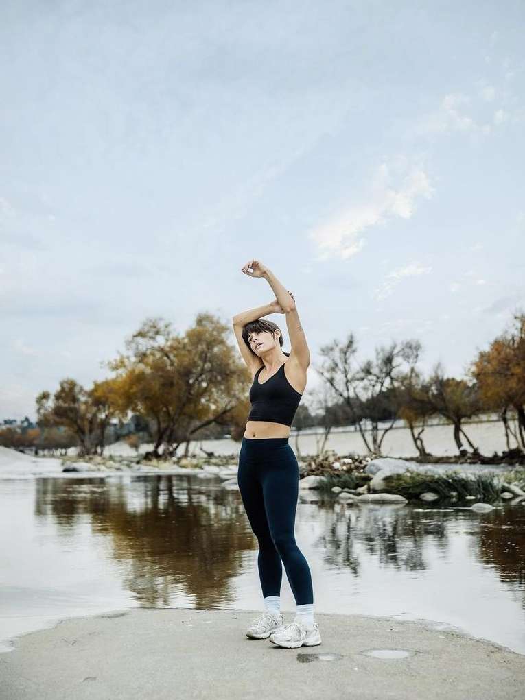 Adult woman in sportswear stretches arms overhead next to a river, trees, and cloudy skies during a workout session outdoors.