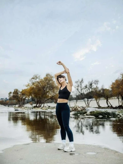 Adult woman in sportswear stretches arms overhead next to a river, trees, and cloudy skies during a workout session outdoors.