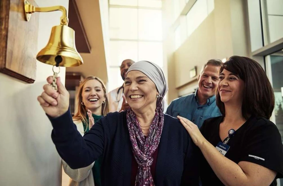 Woman rings celebratory bell surrounded by medical staff and family in a hospital hallway, celebrating recovery milestone after treatment.