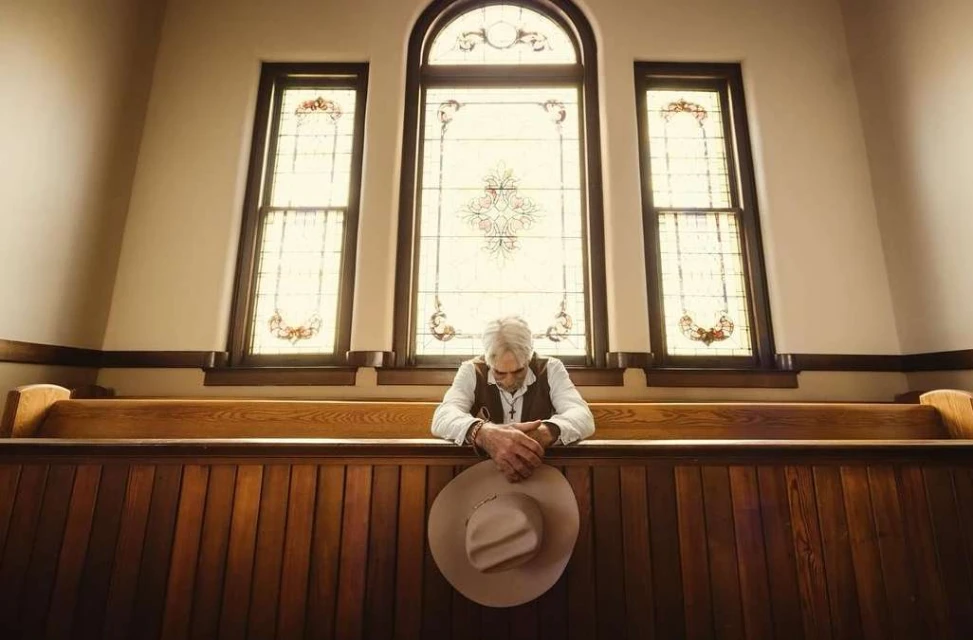 Mature adult man sits in a church pew with his hands clasped together in prayer and a beige cowboy hat resting on the pew's ledge.