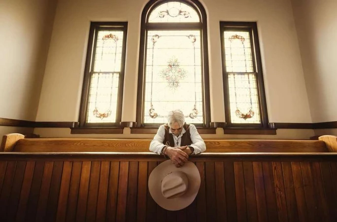 Mature adult man sits in a church pew with his hands clasped together in prayer and a beige cowboy hat resting on the pew's ledge.