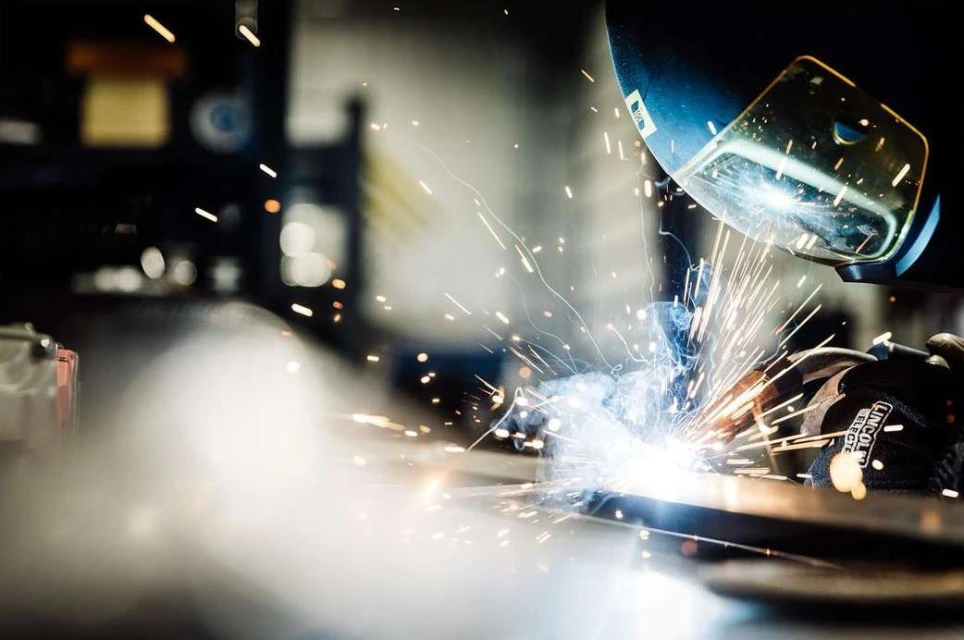 Adult worker welding metal with protective gear, sparks flying in a dark industrial setting, with a blurred background.