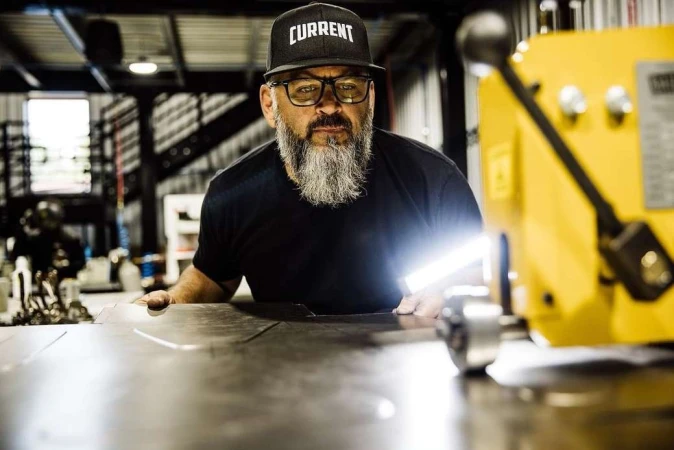 Adult male wearing a 'Current' cap works at a metal fabrication shop, using a yellow metal rolling machine with bright light reflecting.