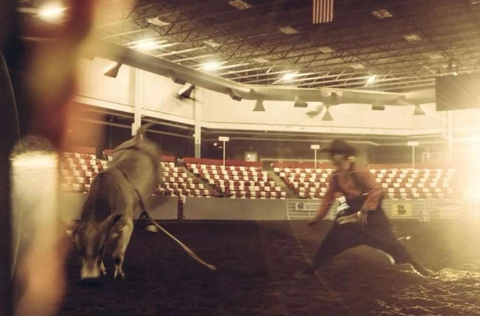 Cowboy faces a bull in an indoor rodeo arena with spectator seating, under bright lighting and an American flag hanging.