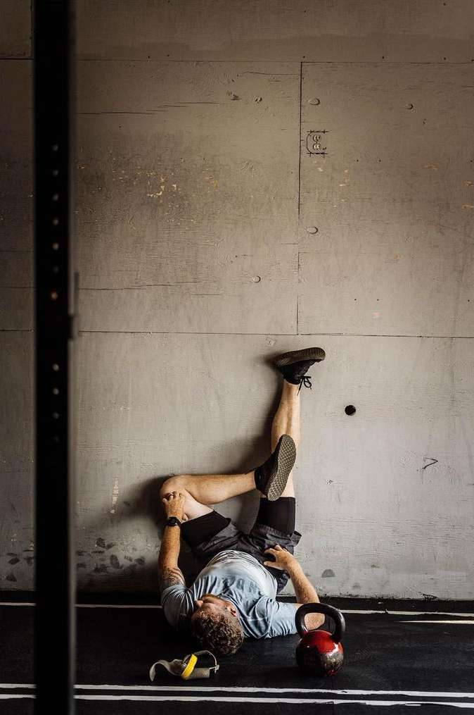 Man lies on a gym floor with his legs resting against a concrete wall in a fitness pose alongside gym equipment and training bands.