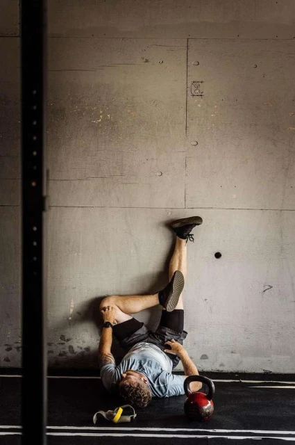 Man lies on a gym floor with his legs resting against a concrete wall in a fitness pose alongside gym equipment and training bands.