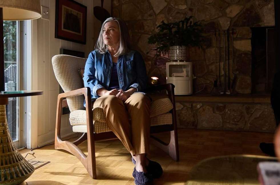 Mature woman with gray hair sitting in an armchair in a wood-paneled room by a window, wearing a blue denim jacket and brown pants.