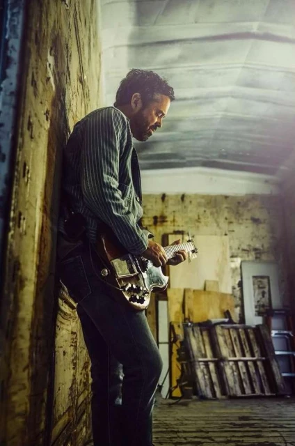 Man plays electric guitar indoors next to grungy wall in jeans and pinstripe jacket with natural light and musical instrument.