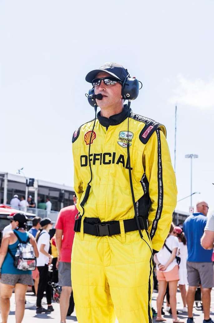 Man in yellow racing suit stands in an outdoor venue, wearing a headset and cap, surrounded by a crowd of people in the background.