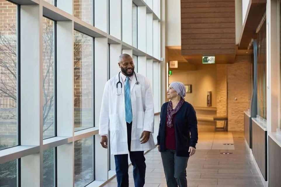 Healthcare scene featuring a smiling African American doctor walking with a female patient in a hospital hallway with large windows.