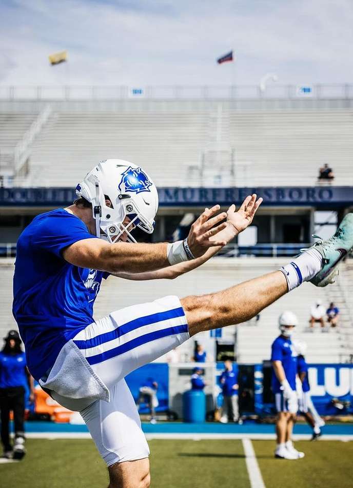 American football kicker in mid-kick on a sunny day with stadium seating in the background and other players on the field.