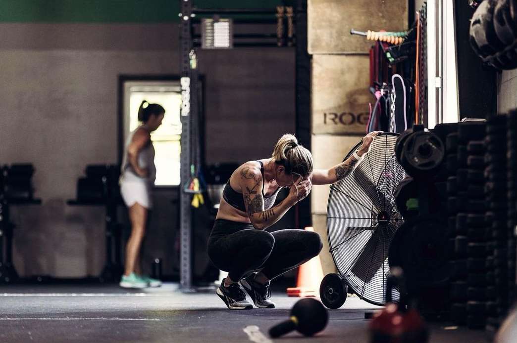 A woman in workout gear squats in a gym with a large fan, showing signs of exertion while another woman stands in the background.