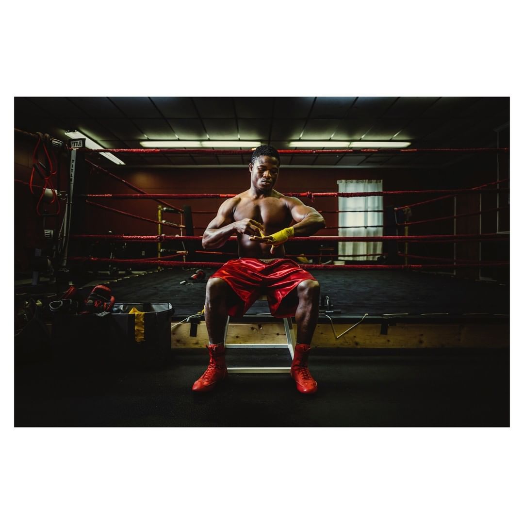 Muscular man wrapping his hands with yellow boxing wraps in a boxing gym, wearing red shorts and boxing shoes, sitting on a wooden stool.