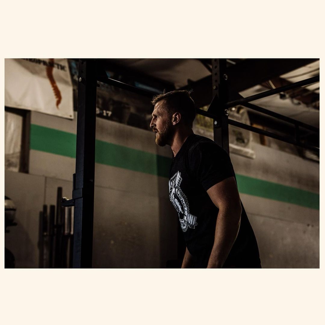 An adult male athlete in a black t-shirt prepares to lift weights at a dimly lit indoor gym with a metal weight rack visible.