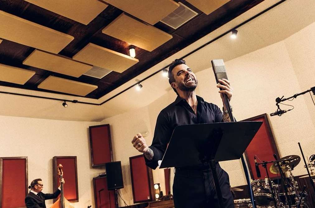 Male singer performing into a vintage microphone in a music studio with musicians and instruments in the background under warm lighting.