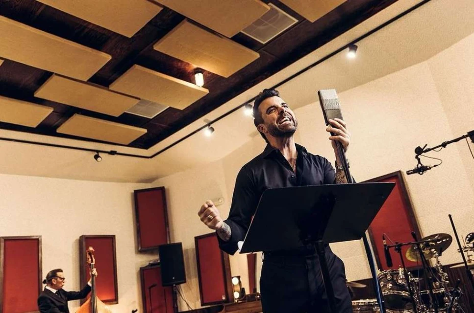 Male singer performing into a vintage microphone in a music studio with musicians and instruments in the background under warm lighting.