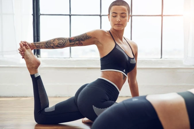 A woman with a buzzcut and tattoos doing a yoga pose in a bright studio wearing black athletic wear.
