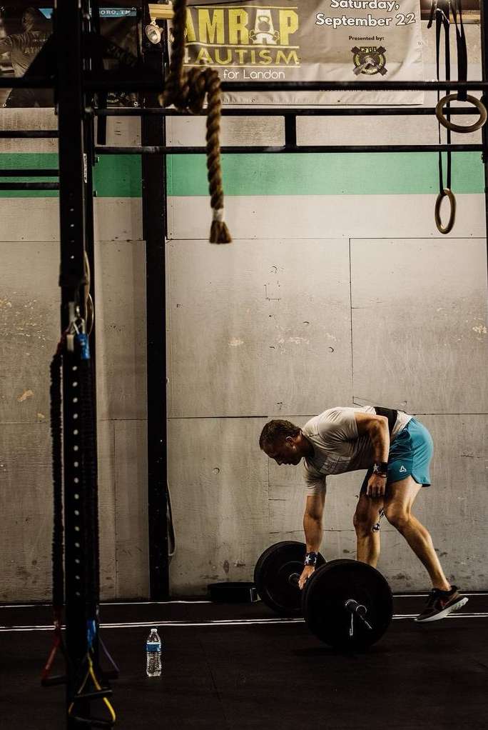 Man in gym prepares to lift barbell, wearing athletic gear and weightlifting belt, with a water bottle nearby, in indoor gym setting.