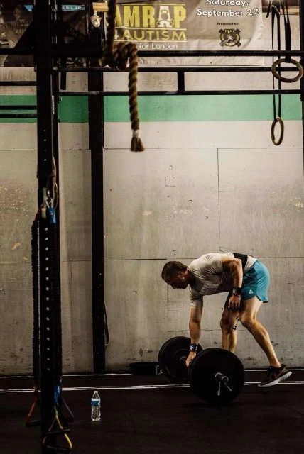 Man in gym prepares to lift barbell, wearing athletic gear and weightlifting belt, with a water bottle nearby, in indoor gym setting.
