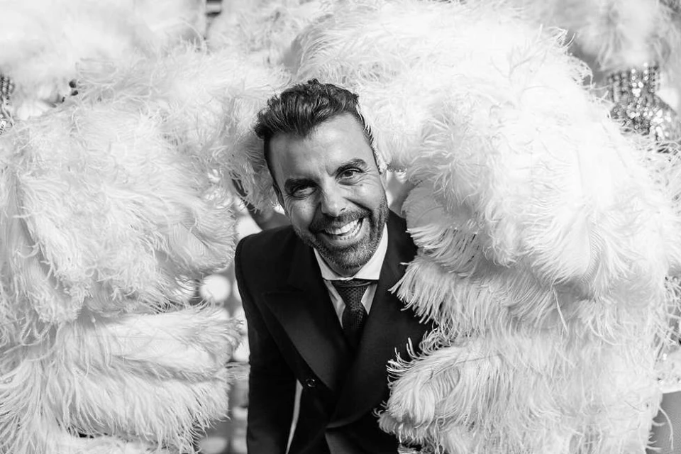 A smiling adult male in a dark suit poses in front of large white feather decorations in a black and white portrait.
