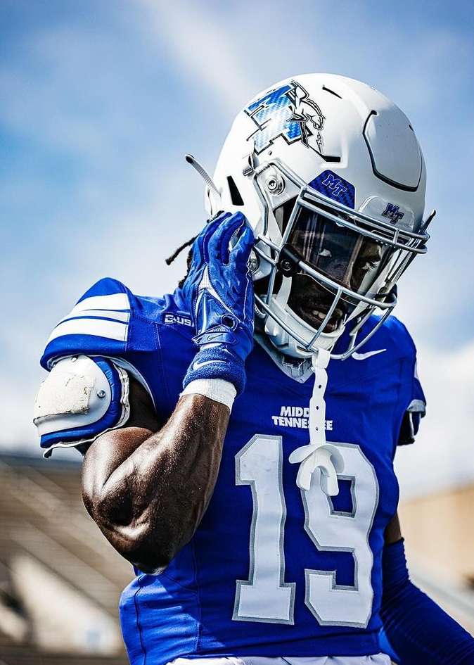 Football player in blue and white uniform wearing a helmet, looks downward against a blue sky background on a sunny day outdoors.