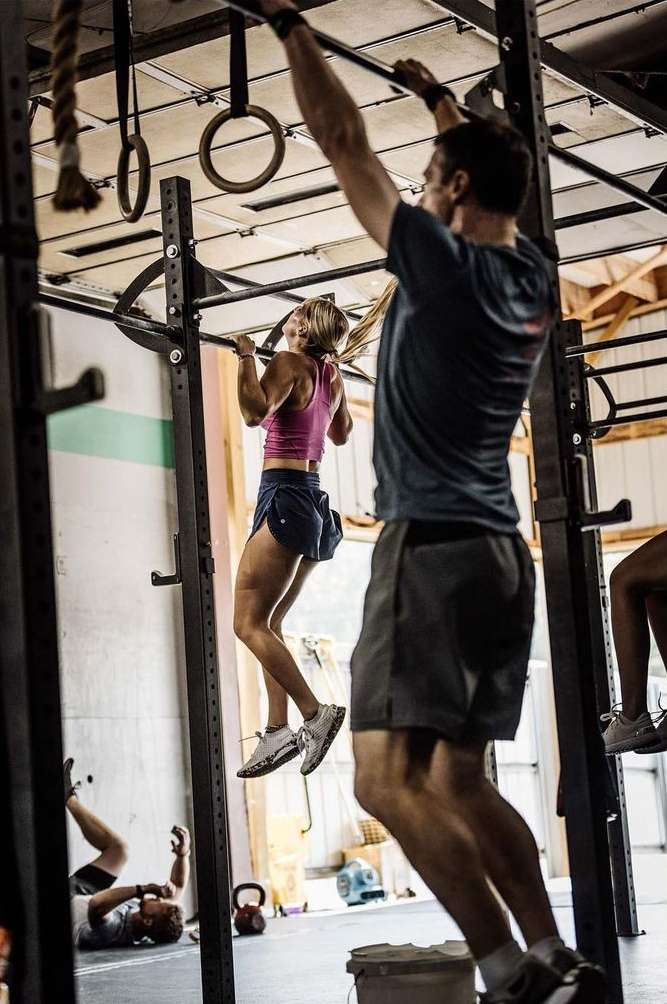 A woman in athletic wear does a pull-up on a metal bar in a gym with a man standing nearby, other people in the background.