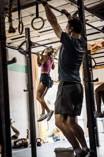 A woman in athletic wear does a pull-up on a metal bar in a gym with a man standing nearby, other people in the background.