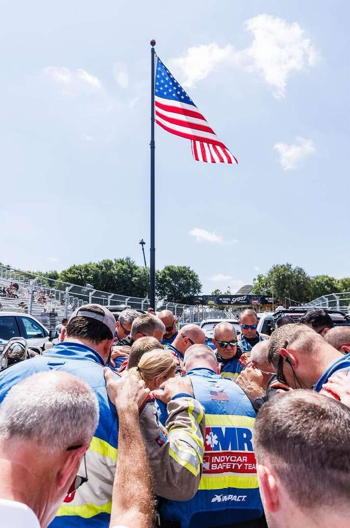 Group of people huddles together under an American flag during the daytime, outdoors. They appear to be praying before a race.