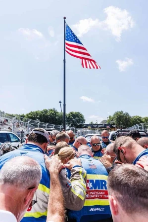Group of people huddles together under an American flag during the daytime, outdoors. They appear to be praying before a race.