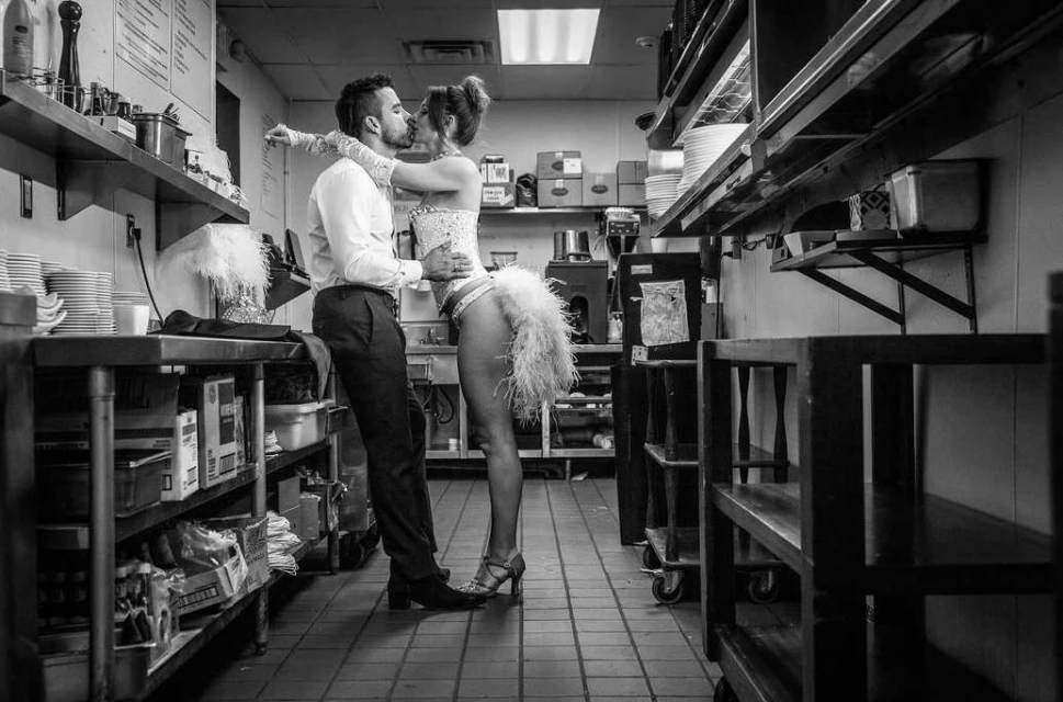 Monochrome shot of a Caucasian couple embracing and kissing in a restaurant kitchen, with metal shelves and equipment in the background.