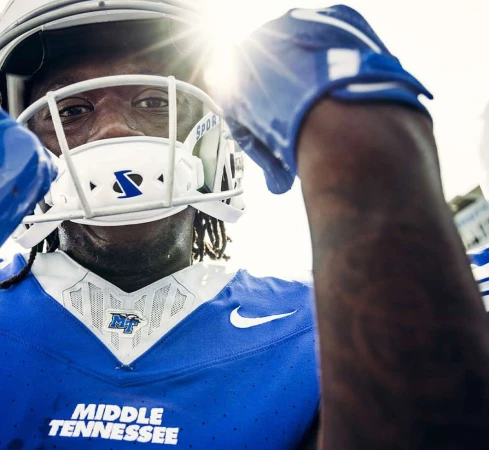 Close-up of an American football player wearing a white helmet and blue jersey, bathed in bright sunlight on the field.