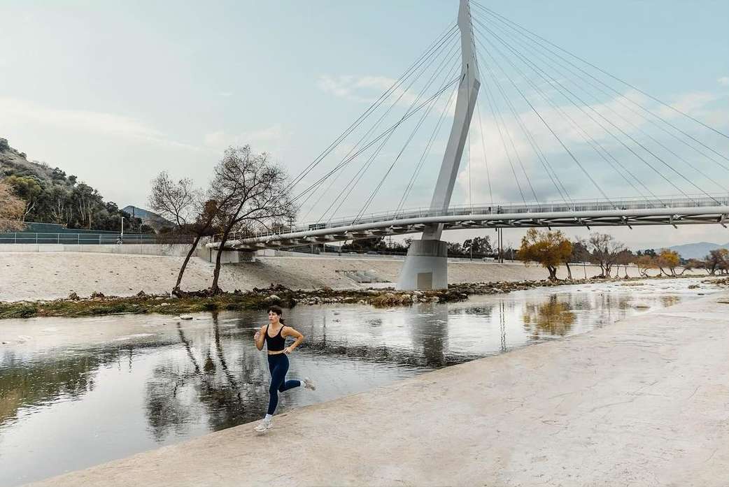 A woman in sportswear is running along a concrete path next to a river; a modern bridge spans the background, fitness, athletics.