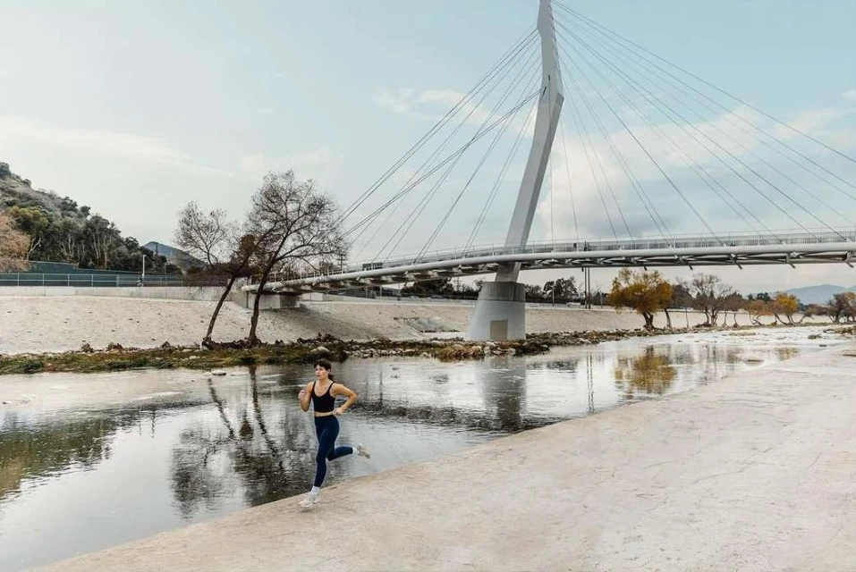 A woman in sportswear is running along a concrete path next to a river; a modern bridge spans the background, fitness, athletics.