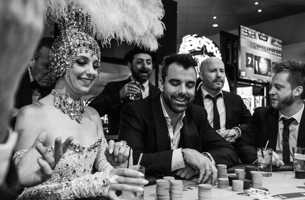 Black and white casino scene with six adults around a poker table. Woman in feathered headpiece smiles with other players during a game.
