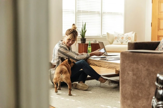 Woman seated on living room floor holding a smartphone with a french bulldog standing nearby by Fab Fernandez
